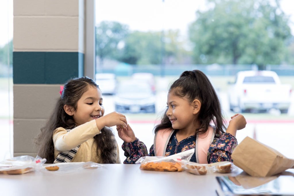 The two young twin sisters share lunch with each other in the elementary school cafeteria.