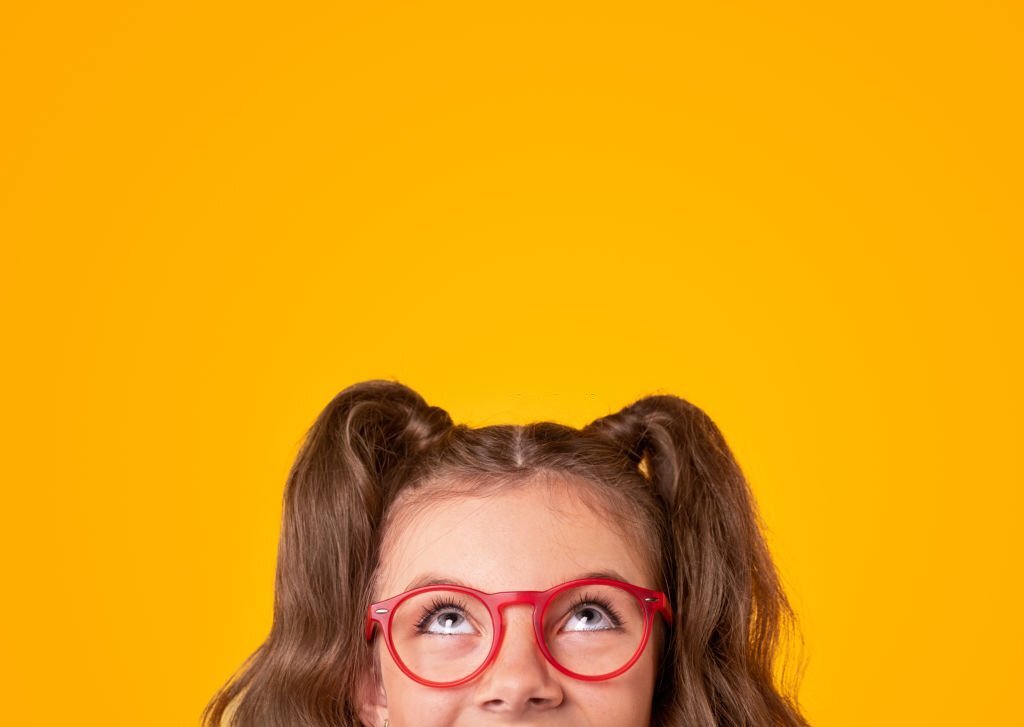 Positive teen girl with ponytails wearing stylish red framed eyeglasses looking up on yellow background with empty space
