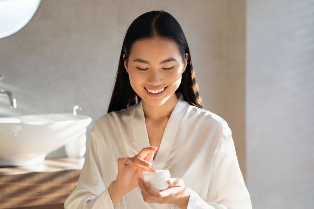 Smiling attractive young asian lady in white bathrobe holding jar with beauty product, enjoying morning routine for beautiful face skin, standing in modern bathroom, sun flare, copy space