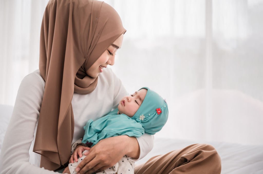 Happy muslim mother holding adorable little baby daughter wearing hijab in her arms on white bed in bedroom. Arab young mom wearing a head scarf, and she's sitting on the bed while holding her baby.