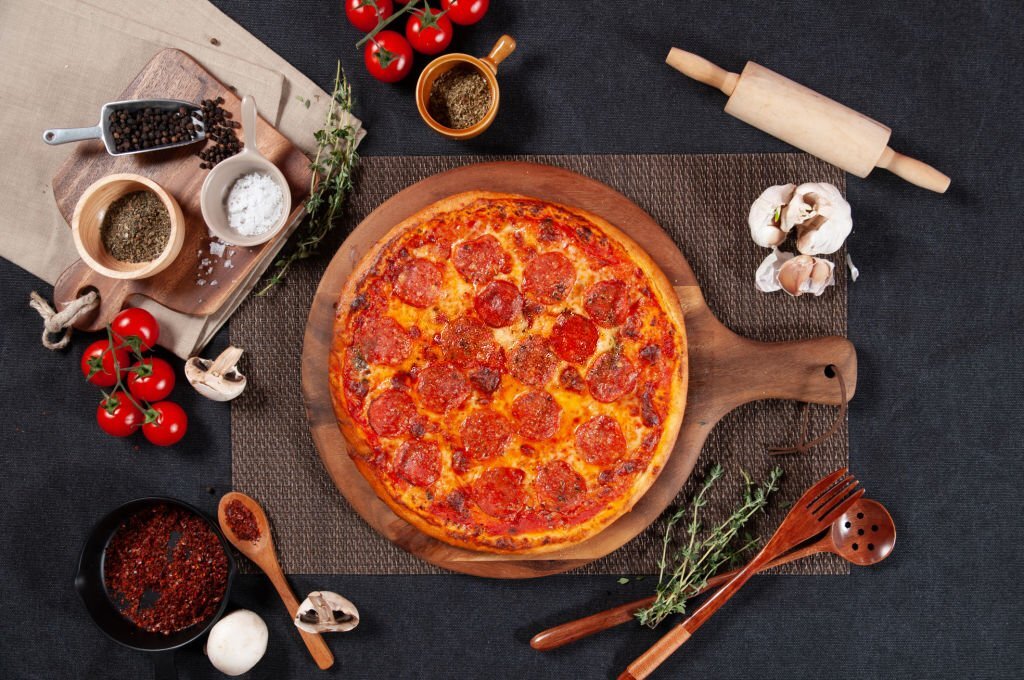 Traditional Pepperoni Pizza with raw cherry tomato, black pepper, garlic, and mushroom isolated on wooden cutting board on dark background top view of fastfood