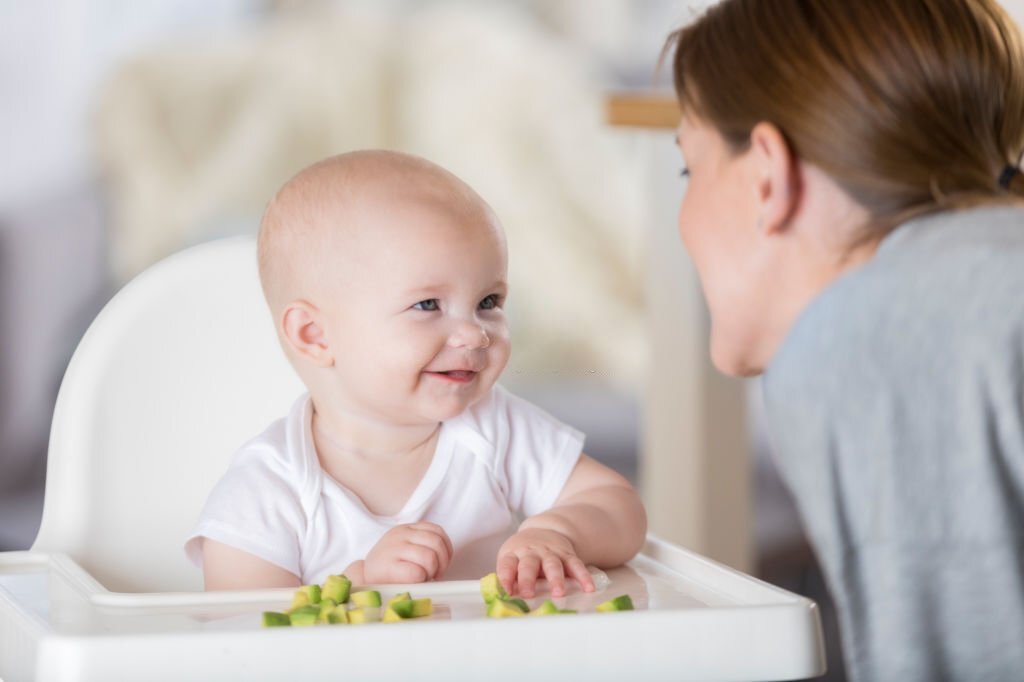 An adorable baby sits in a high chair and smiles at her mother as she reaches for finger food on the tray.