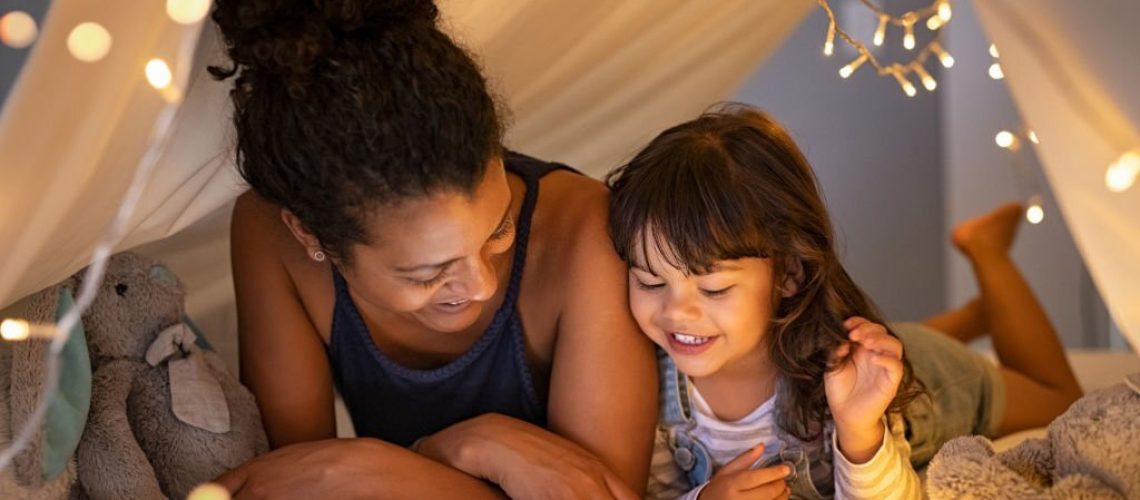 African mother and cute smiling girl using digital tablet while lying in illuminted tent in kid bedroom. Cheerful ethnic woman and lovely daughter on video call under a cozy hut. Lovely little girl with mom watching cartoon on digital tablet in bedroom.