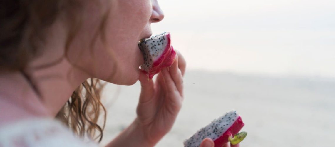 Close-up shot of a young caucasian woman eating dragon fruit on the beach.