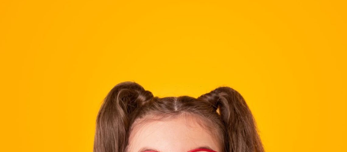 Positive teen girl with ponytails wearing stylish red framed eyeglasses looking up on yellow background with empty space