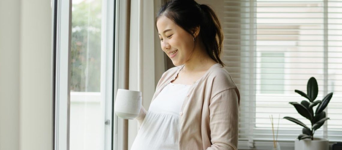 pregnancy, drinks, rest, people and expectation concept - close up of happy pregnant woman with cup drinking tea looking through window at home