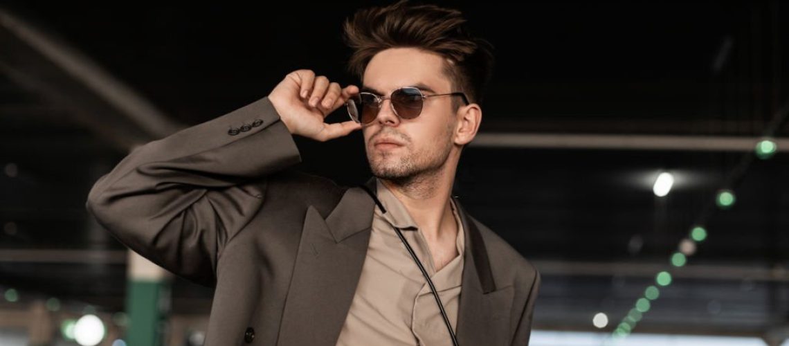 Handsome young business man with hairstyle in fashionable clothes look straightens sunglasses and walks on the street. Male casual style and fashion