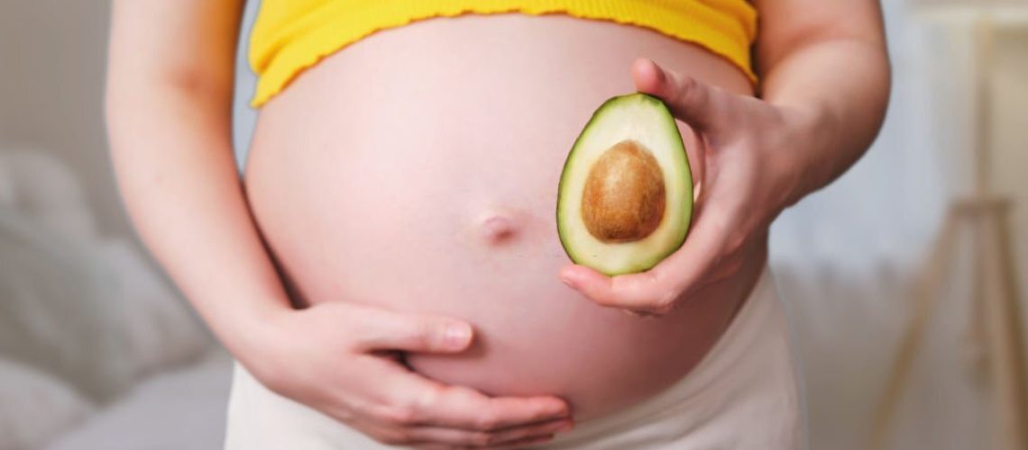 Avocado with a bone in the hands of a pregnant woman, home living room