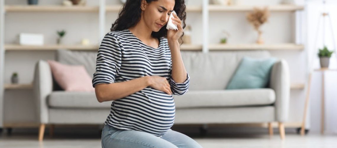 Pregnancy hormonal changes concept. Young curly long-haired pregnant woman feeling down, sitting on floor in cozy living room and crying, using napkins, staying alone at home, free space