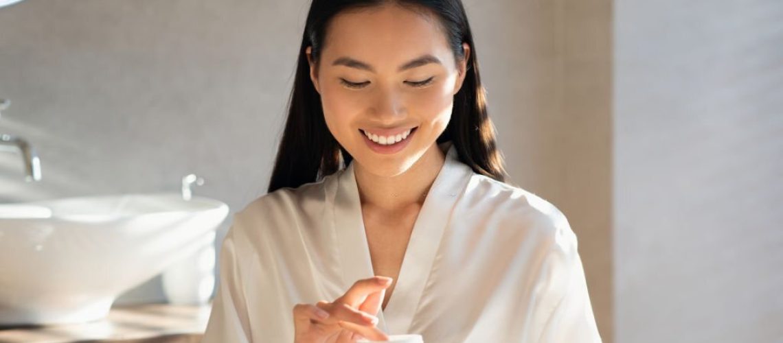 Smiling attractive young asian lady in white bathrobe holding jar with beauty product, enjoying morning routine for beautiful face skin, standing in modern bathroom, sun flare, copy space