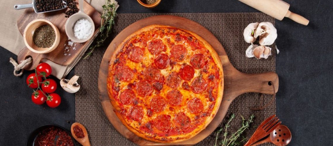 Traditional Pepperoni Pizza with raw cherry tomato, black pepper, garlic, and mushroom isolated on wooden cutting board on dark background top view of fastfood