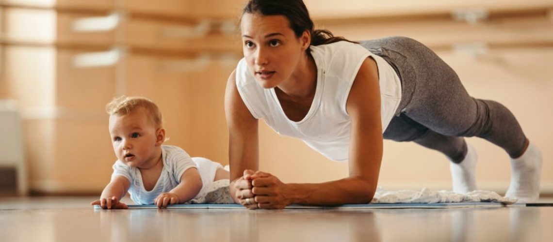 Athletic mother exercising in plank position while being with her baby in a health club. Copy space.