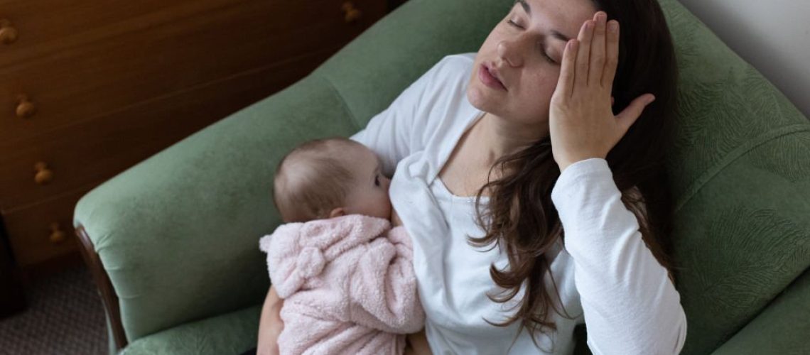 Mother experiences headache while breastfeeding her daughter. Mother sit in the armchair and holds her daughter in her arms while breastfeeding. Baby one year old. The mother takes care of her daughter on maternity leave.