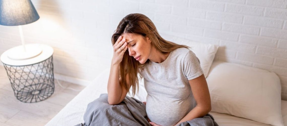 Young pregnant woman having headache. She is resting on bed in the bedroom and feeling unwell.