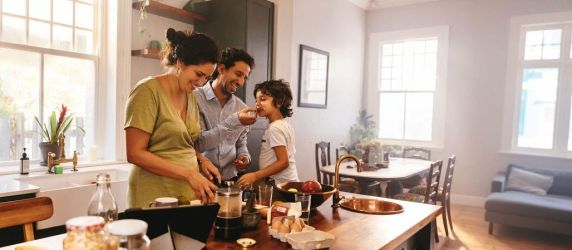 Playful dad feeding his son a slice of bread while his wife prepares breakfast. Family of three having fun together in the kitchen. Mom and dad spending quality time with their son.