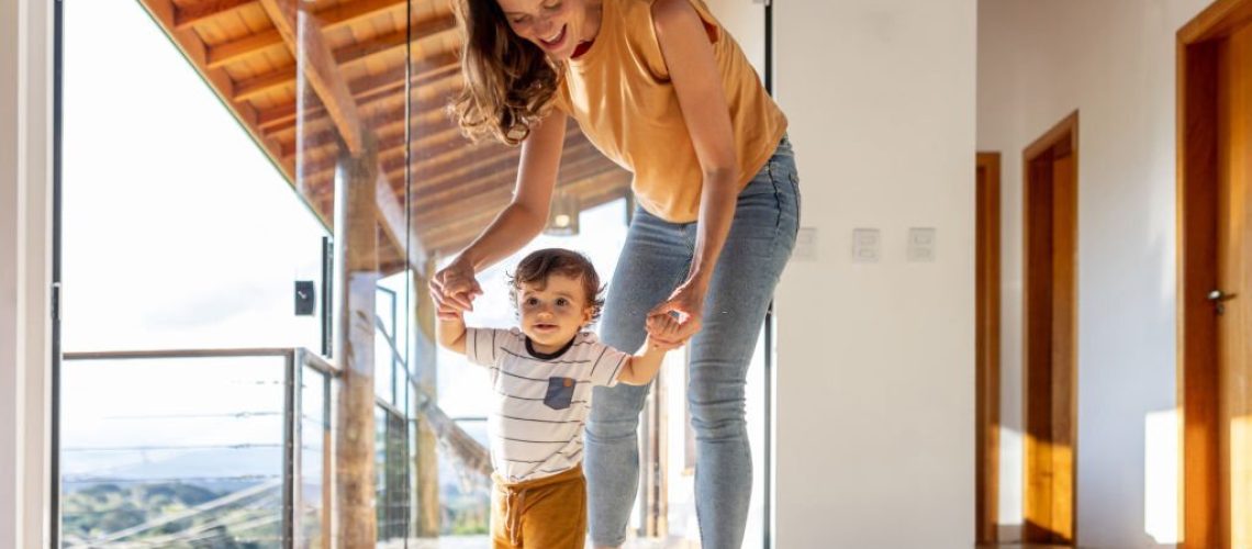 Happy toddler at home learning how to walk with the help of his mother - first steps concepts
